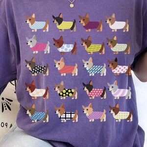 May include: White, purple, and green t-shirts featuring a repeating pattern of corgi dogs wearing various outfits. The shirts have the text "GIFTS FOR CORGI LOVERS" at the bottom. The corgis are wearing sweaters with patterns like stripes, polka dots, and plaid.