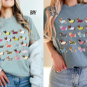 May include: Two t-shirts, one in bay and one in blue jean, each featuring a repeating pattern of dachshunds wearing various patterned sweaters. The shirts are short-sleeved and made of a soft material.