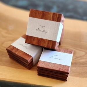 May include: Set of six square wooden coasters with a natural wood finish. Each coaster has a white label with the text "HANDCRAFTED BY L. Treublay".