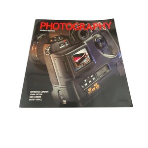Puede incluir: Una cámara negra y plateada con una lente roja y una pantalla en blanco y negro. La cámara está en la portada de un libro titulado "Photography, Seventh Edition" de Barbara London, John Upton, Ken Kobré y Betsy Brill.