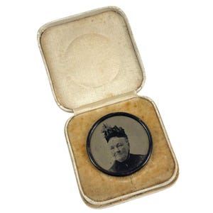 Puede incluir: Un retrato en blanco y negro de una mujer con sombrero se muestra en una pequeña caja cuadrada blanca con forro de terciopelo.