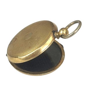 Puede incluir: Un medallón de oro con un interior negro. El medallón tiene un diseño circular y un pequeño bucle en la parte superior.