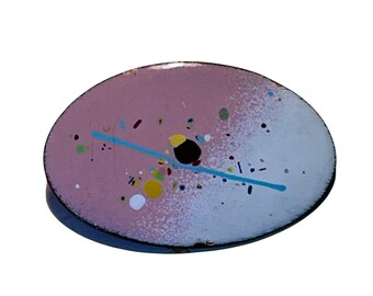 May include: Oval enamel pin with a pink and white surface. The pin has a blue line and colourful paint splatters. The pin has a black border.