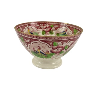 以下が含まれることがあります： 金色の縁取りと白い台座のある、ピンクと緑のヴィンテージのフラワーボウル。ボウルには、鳥と花が繰り返し描かれた模様があります。