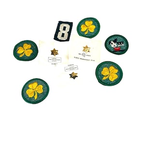 Puede incluir: Una colección de parches y pines de Girl Scouts, que incluye un parche de trébol verde con un trébol amarillo, una estrella de membresía dorada y un parche con el número 8.
