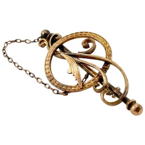 May include: A gold-toned brooch with a delicate, ornate design featuring a circle with a stylized leaf inside. The brooch has a chain attached to the top.