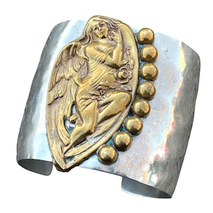 May include: A silver cuff bracelet with a brass angel figure and a row of brass beads on one side.