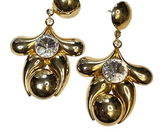 May include: A pair of gold-toned earrings with a unique design. Each earring features a large, clear, round gemstone at the center, surrounded by a stylized, petal-like shape. A smaller, round gold ball hangs below the gemstone.