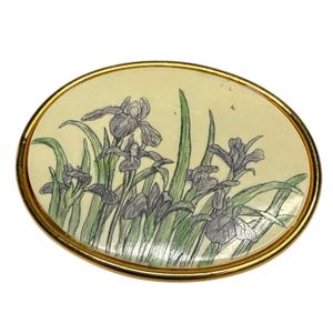 Könnte beinhalten: Ovale Brosche mit goldfarbenem Rahmen. Die Brosche zeigt ein gemaltes Design aus lila Iris und grünen Blättern auf cremefarbenem Hintergrund. Das florale Design ist detailliert und künstlerisch und schafft eine Vintage-Ästhetik.
