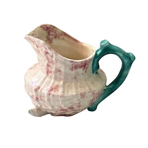 May include: A ceramic pitcher with a white base and a pattern of red spots. The handle is a teal green color. The pitcher has a unique shape and design, with a spout for pouring. The pitcher is likely used for serving beverages.