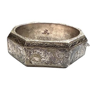 Puede incluir: Un brazalete vintage articulado de plata de ley. El brazalete tiene forma octogonal con grabados florales y de volutas ornamentados. El interior es liso y el cierre es una pequeña lengüeta. El brazalete es de color plateado.