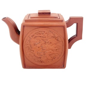 May include: A square terracotta teapot with a dragon design on the front. The teapot has a lid and a handle.