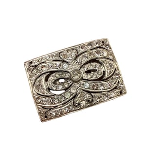 May include: A rectangular, silver-toned brooch with a bow design. The brooch is encrusted with numerous small, clear rhinestones, and a larger, central rhinestone. The edges of the brooch have a decorative, etched pattern.