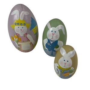 May include: Three wooden Easter eggs decorated with painted bunnies. The largest egg is purple with a bunny wearing a pink dress and holding flowers. The middle egg is green with a bunny wearing a blue vest and holding a carrot. The smallest egg is yellow with a bunny wearing a blue and yellow scarf and holding a carrot.