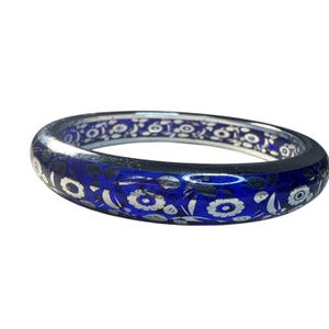 May include: A blue bangle bracelet with a floral design. The bracelet has a silver-colored metal rim and a dark blue band with white floral patterns. The flowers and leaves are intricately detailed, creating a delicate and elegant look.