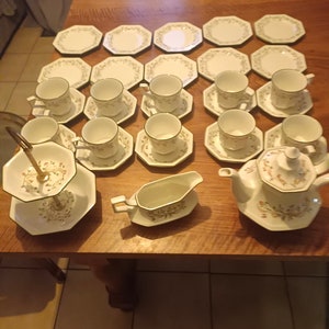 Op de afbeelding: Een wit theeservies met een groen bloemenpatroon. De set bevat een theepot, een roomkan, een suikerpot en 12 kopjes en schoteltjes. De set is op een houten tafel gerangschikt.