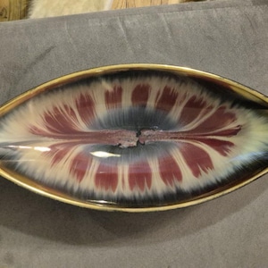 Könnte beinhalten: Eine ovale Keramikschale mit einzigartigem Design. Das Innere zeigt ein zentrales Muster aus roten und weißen Strahlenlinien vor einem blauen und schwarzen Hintergrund. Der Rand ist goldfarben.