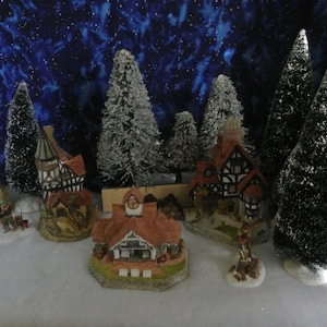 Può includere: Una scena di villaggio natalizio in miniatura con case, alberi e figure ricoperte di neve. Il villaggio è situato su uno sfondo blu scuro con stelle bianche.