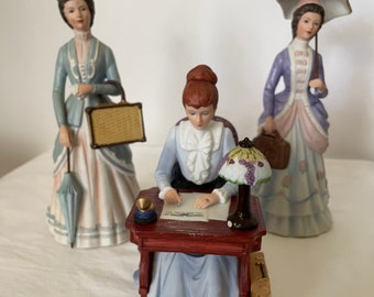 May include: Three porcelain figurines of women in Victorian era clothing. One woman is seated at a desk writing a letter, another is holding an umbrella and a handbag, and the third is holding a suitcase and an umbrella.