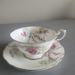 Könnte beinhalten: Weißes Tee-Set mit Blumen- und Vogelmuster. Die Tasse und Untertasse haben einen goldenen Rand. Die Tasse zeigt zwei Vögel und rosa Rosen. Die Untertasse hat einen gewellten Rand.