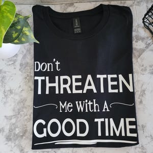 Può includere: T-shirt nero con la scritta bianca "Don't THREATEN Me With A GOOD TIME". La maglietta è piegata e si trova su una superficie grigia e bianca. Una pianta verde è visibile nell'angolo in alto a sinistra.