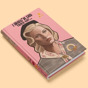 May include: A pink journal with the text "I Should've Said Therese Wait..." and the name "Carol" on the spine. The cover features an illustration of a woman with blonde hair and a black hat. Gold stars are also on the cover.