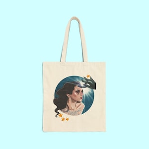 May include: Cream-colored tote bag featuring a circular illustration of a woman with long dark hair, a green hand, and radiating light. The text "MAGINAL DISK" is below the woman. Gold stars are scattered around the illustration.