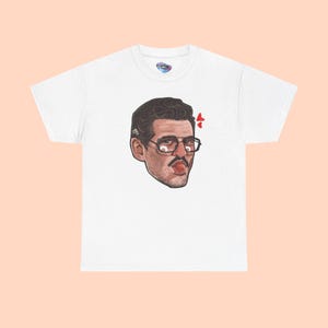 May include: White t-shirt featuring a cartoon illustration of a man with glasses, a mustache, and his tongue sticking out. The graphic includes red heart accents. The shirt has a crew neck and short sleeves.