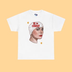 May include: White t-shirt featuring a graphic of a person wearing a white headscarf with the text "BE THE COWBOY" in red. The person has red lipstick and is surrounded by yellow stars.