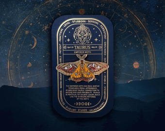 Op de afbeelding: Een gouden emaille pin met een mot met rode, gele en zwarte vleugels. De pin is omgeven door een ronde lijst met de woorden "Taurus Emperor Moth" en de data "April 20" en "May 20". De lijst is versierd met sterren en de woorden "Loyal, Stur, Sensueel, Volhardend, Geaard, Diep, Stabiel, Standvastig, Vastberaden, Warmhartig, Geduldig, Praktisch, Artistiek, Zachtaardig, Waardevol, Betrouwbaar".
