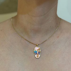 May include: Gold-toned necklace with a pendant featuring an abstract face design. The pendant has a pink background with blue, orange, and white accents. The necklace is worn around the neck.
