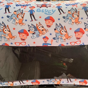 Puede incluir: Una capota de cochecito con un patr&oacute;n repetido de los personajes de dibujos animados Bluey y Bingo, Blippi y otros gr&aacute;ficos sobre un fondo azul claro. Tiene una ventana de malla negra.