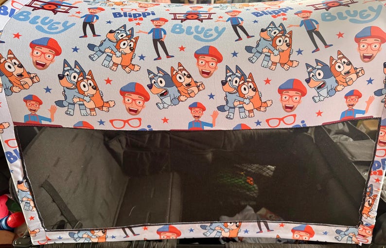 Puede incluir: Una capota de cochecito con un patr&oacute;n repetido de los personajes de dibujos animados Bluey y Bingo, Blippi y otros gr&aacute;ficos sobre un fondo azul claro. Tiene una ventana de malla negra.