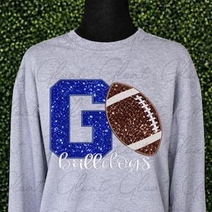 May include: A light gray long-sleeve sweatshirt featuring a large blue glitter letter G next to a brown glitter football graphic. Below the graphics, the word bulldogs is printed in white script lettering on the front of the garment.