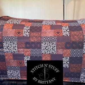 May include: A patchwork fabric seat cover with a brown, white, and red color scheme. The fabric features a repeating pattern of geometric shapes and a logo with a stylized "W" inside a circle. The text "STITCH N' STUFF BY BRITTANY" is printed on the bottom of the cover.