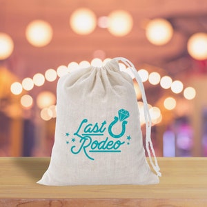 May include: A white drawstring bag with the text "Last Rodeo" and a turquoise horseshoe with a diamond on top. The bag is sitting on a wooden surface.