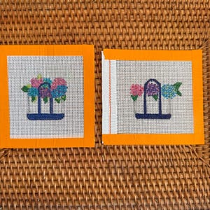 Puede incluir: Dos lienzos cuadrados de punto de aguja con bordes naranjas. Cada lienzo presenta un dise&ntilde;o floral en rosa, azul y verde, sobre un fondo blanco. Los arreglos florales est&aacute;n en una estructura en forma de jarr&oacute;n azul oscuro. Los lienzos se muestran sobre una superficie de mimbre tejido.