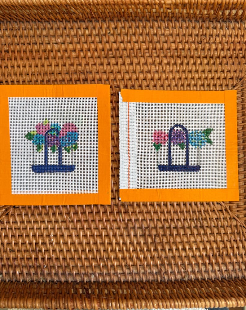 Puede incluir: Dos lienzos cuadrados de punto de aguja con bordes naranjas. Cada lienzo presenta un dise&ntilde;o floral en rosa, azul y verde, sobre un fondo blanco. Los arreglos florales est&aacute;n en una estructura en forma de jarr&oacute;n azul oscuro. Los lienzos se muestran sobre una superficie de mimbre tejido.