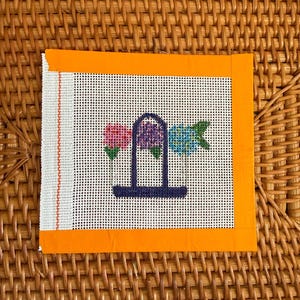 Puede incluir: Lienzo de punto de aguja con dise&ntilde;o floral. El lienzo tiene un fondo blanco con un patr&oacute;n de cuadr&iacute;cula. El dise&ntilde;o incluye tres flores en rosa, morado y azul, sobre una estructura arqueada azul oscuro. El lienzo est&aacute; enmarcado con un borde naranja.