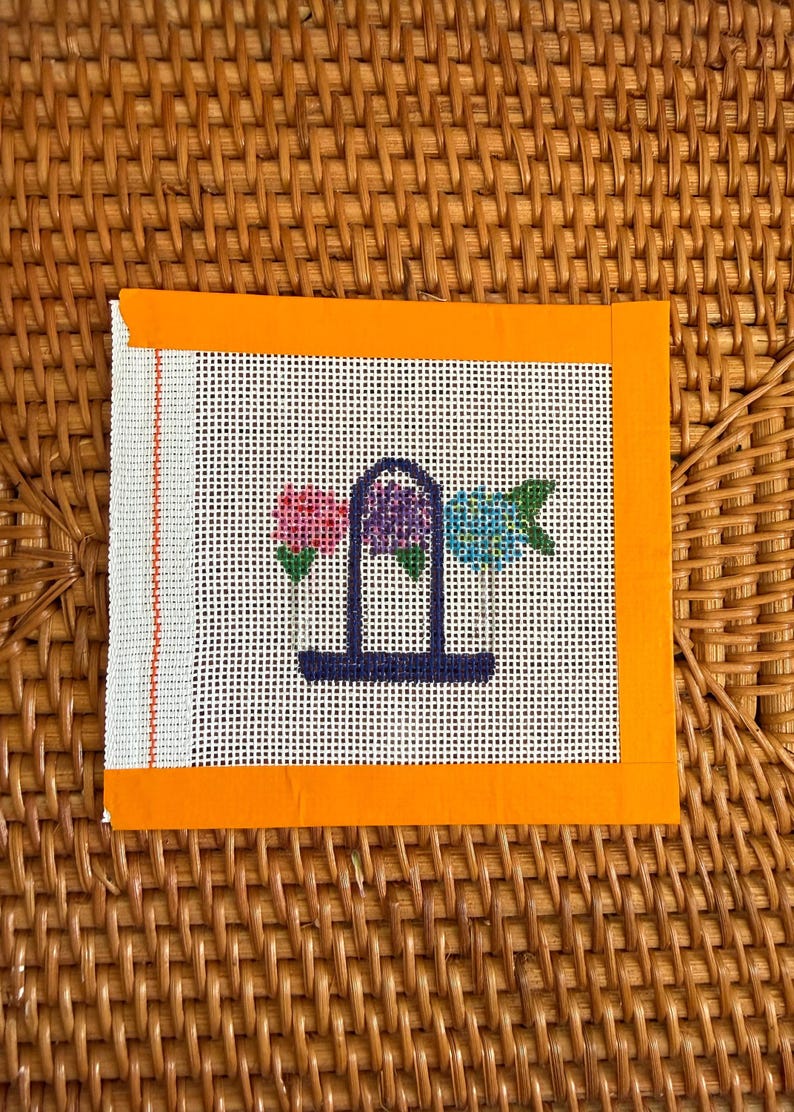 Puede incluir: Lienzo de punto de aguja con dise&ntilde;o floral. El lienzo tiene un fondo blanco con un patr&oacute;n de cuadr&iacute;cula. El dise&ntilde;o incluye tres flores en rosa, morado y azul, sobre una estructura arqueada azul oscuro. El lienzo est&aacute; enmarcado con un borde naranja.