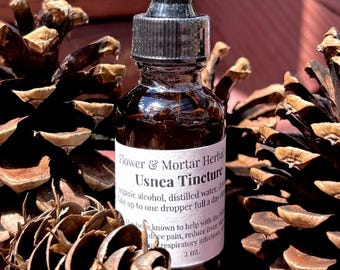 May include: A brown glass bottle of Usnea Tincture with a black dropper, labeled "Flower & Mortar Herbal." The bottle is surrounded by pine cones on a red wooden surface. The label includes text about the product.