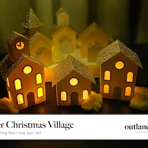 May include: A paper Christmas village with a clock tower and several other houses, all lit up with warm yellow lights. The houses are made of paper and have a glittery finish. The village is sitting on a brown surface.