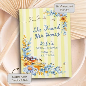 May include: A hardcover lined journal measuring 8 inches by 5.75 inches with a yellow and white striped cover. The text reads She Found Her Honey, Katie's BRIDAL SHOWER, MIAMI, FL, JULY 2026. It features illustrations of blue and yellow flowers and bees.