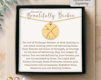 May include: A gold-toned circular pendant necklace featuring a Kintsugi-inspired cracked design. The jewellery is presented in a black box on a card with the text You are so Beautifully Broken and a paragraph explaining the art of Kintsugi.