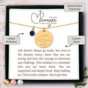 May include: Gold necklace with a circular pendant engraved with a wave design and the name "Rachel". A blue gemstone and the words "You are stronger than the storm" are also visible. The necklace is presented in a black box.