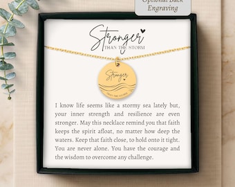 Può includere: Una collana con ciondolo circolare dorato su una catenina sottile, presentata in una scatola nera con un biglietto che riporta il testo Stronger Than The Storm. Il ciondolo è inciso con la parola Stronger, un cuoricino e linee ondulate.