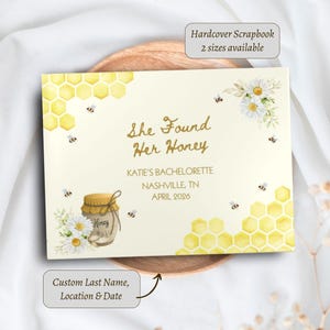 May include: A rectangular hardcover scrapbook with a cream background featuring yellow honeycomb patterns, white daisies, and small bees. The text reads She Found Her Honey, KATIE'S BACHELORETTE, NASHVILLE, TN, APRIL 2026, and includes a honey jar illustration.