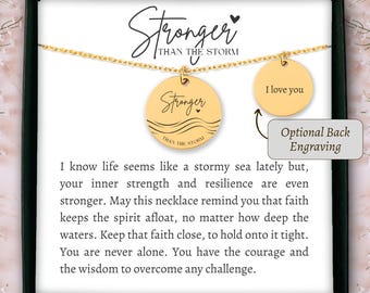 Könnte beinhalten: Goldfarbene Halskette mit zwei runden Anhängern. Einer trägt die Aufschrift "Stronger Than The Storm", der andere "I love you". Die Kette wird auf einer weißen Karte mit inspirierendem Text präsentiert.