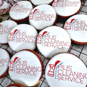 May include: A collection of round cookies with white icing, featuring the text "PILIS CLEANING SERVICE" in red. Each cookie has a red Santa hat graphic and a cleaning spray bottle graphic. The cookies sit on a wire rack.