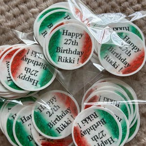 May include: A collection of round stickers with a green, white, and red color scheme, resembling a flag. Each sticker has the text "Happy 27th Birthday Rikki!" printed on it. The stickers are packaged in clear plastic.